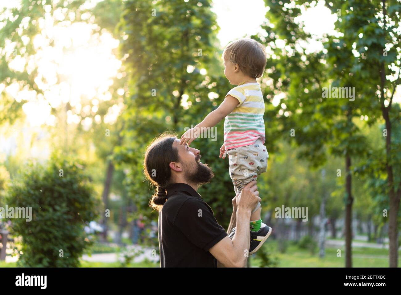 Paternità, paternità, infanzia, cura, estate e tempo libero - il giovane papà con barba e capelli lunghi in t-shirt nera tiene in braccio poco Foto Stock