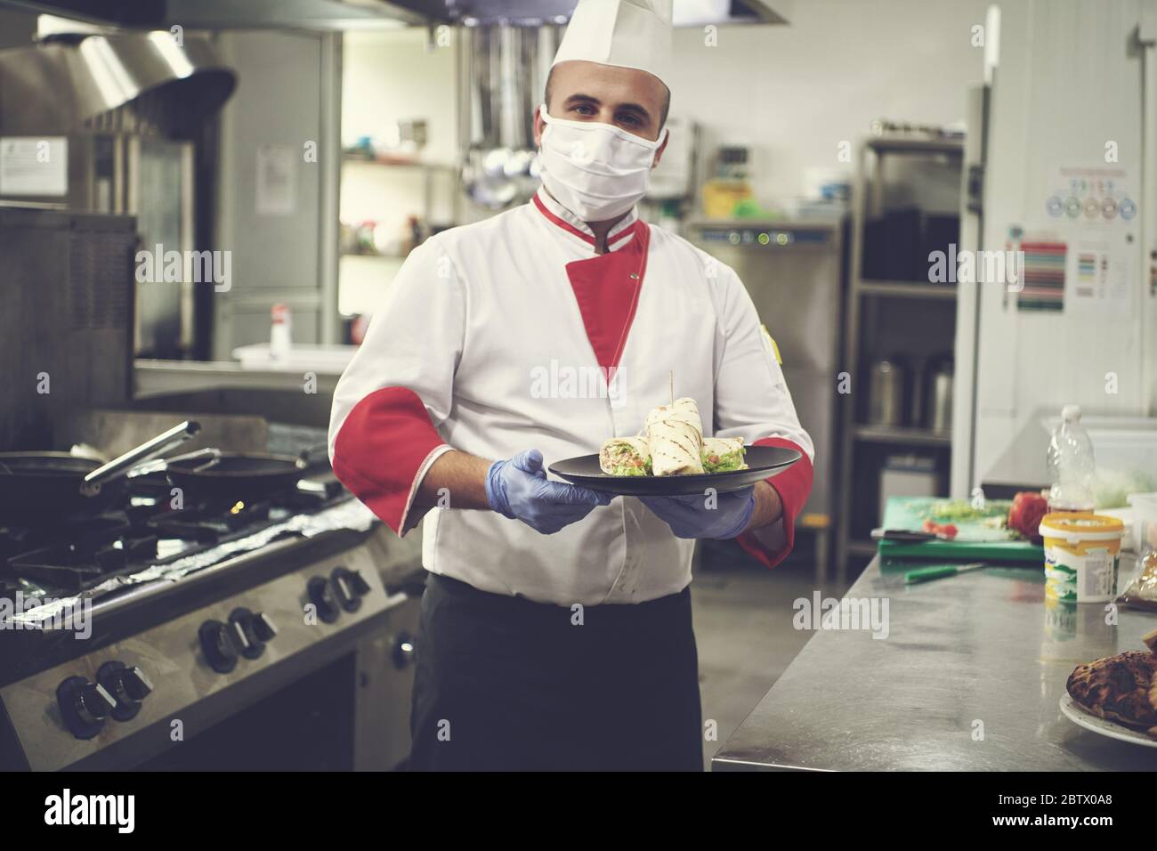 cuoco cuoco che indossa maschera medica protettiva per la protezione dalla malattia di corona virus con piatto di autentico sandwich al ristorante professionale ki Foto Stock