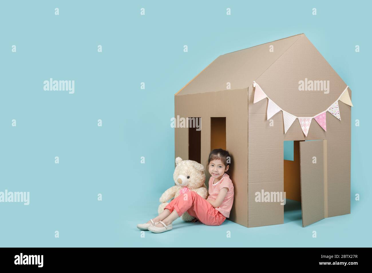 Ragazza bambino asiatica che gioca con la casa di cartone con il suo orsacchiotto isolato su banner blu lungo con spazio di copia per il tuo testo, Creative a casa Foto Stock
