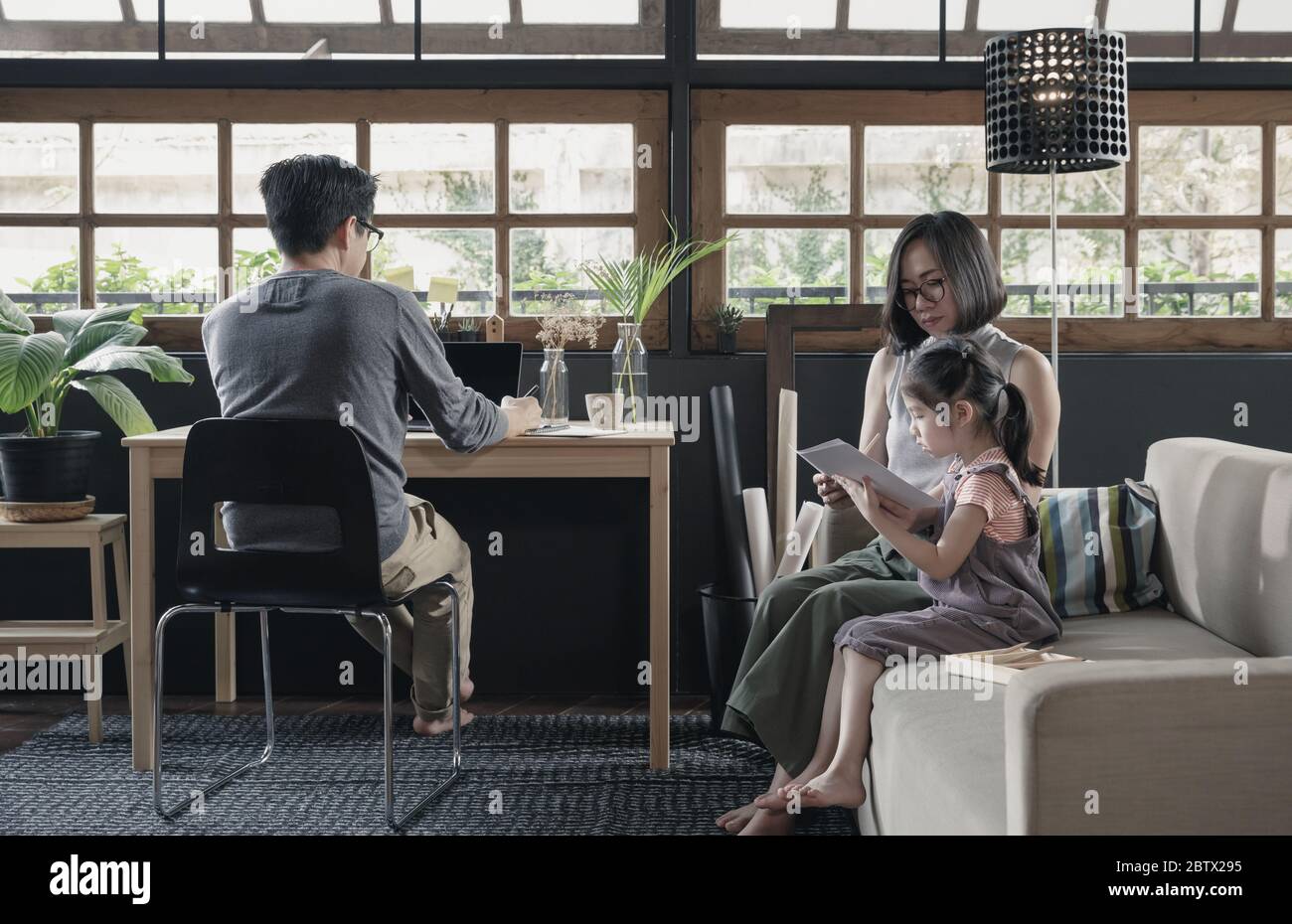 Lavorando da casa di vita padre utilizzando il computer portatile sulla scrivania e bambina imparare con la madre sul divano nel soggiorno, isolamento quarantena d Foto Stock