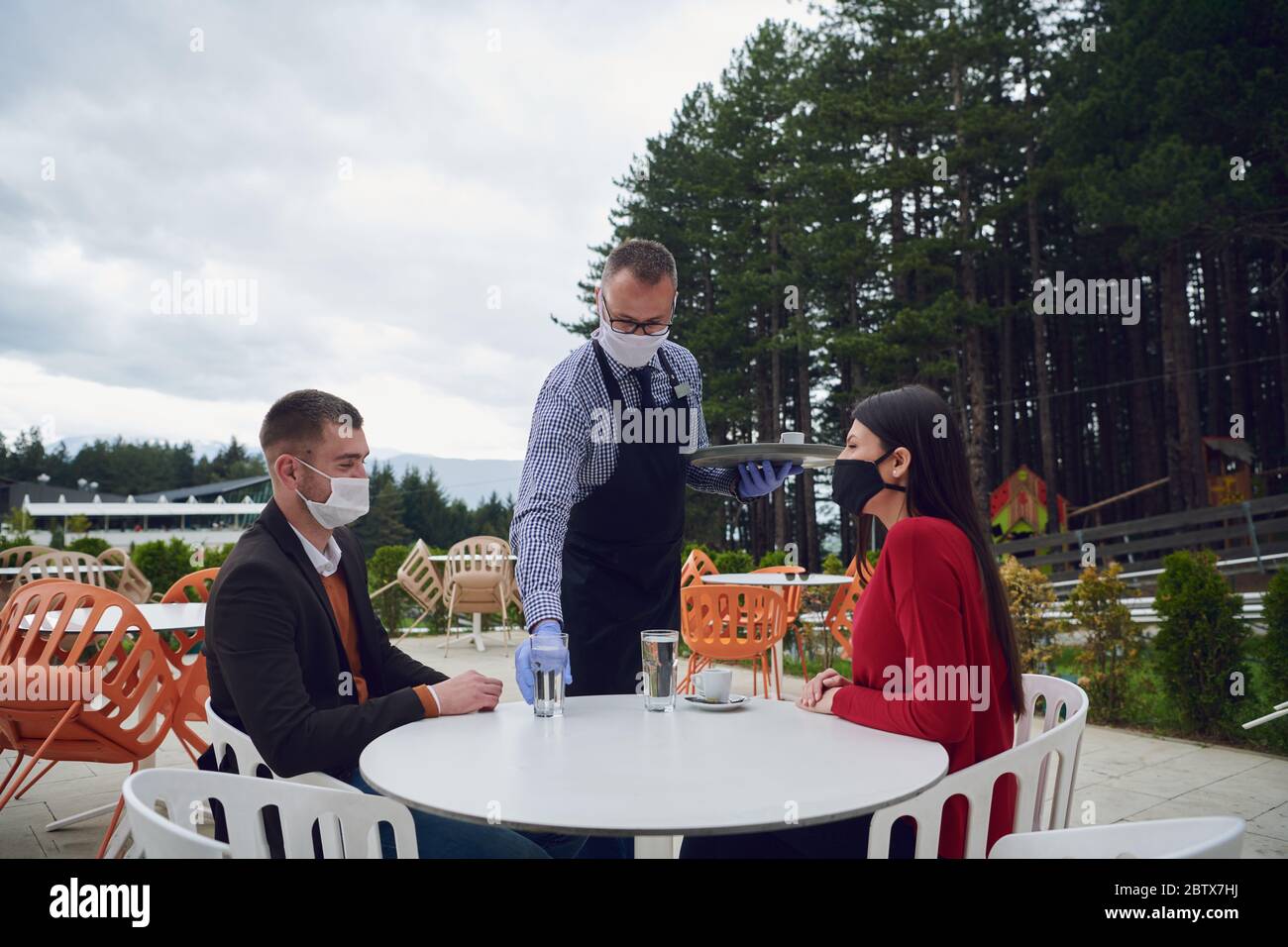 Cameriere con maschera medica protettiva e guanti per servire gli ospiti con caffè in un bar all'aperto o ristorante nuovo concetto normale riapertura dopo quara Foto Stock