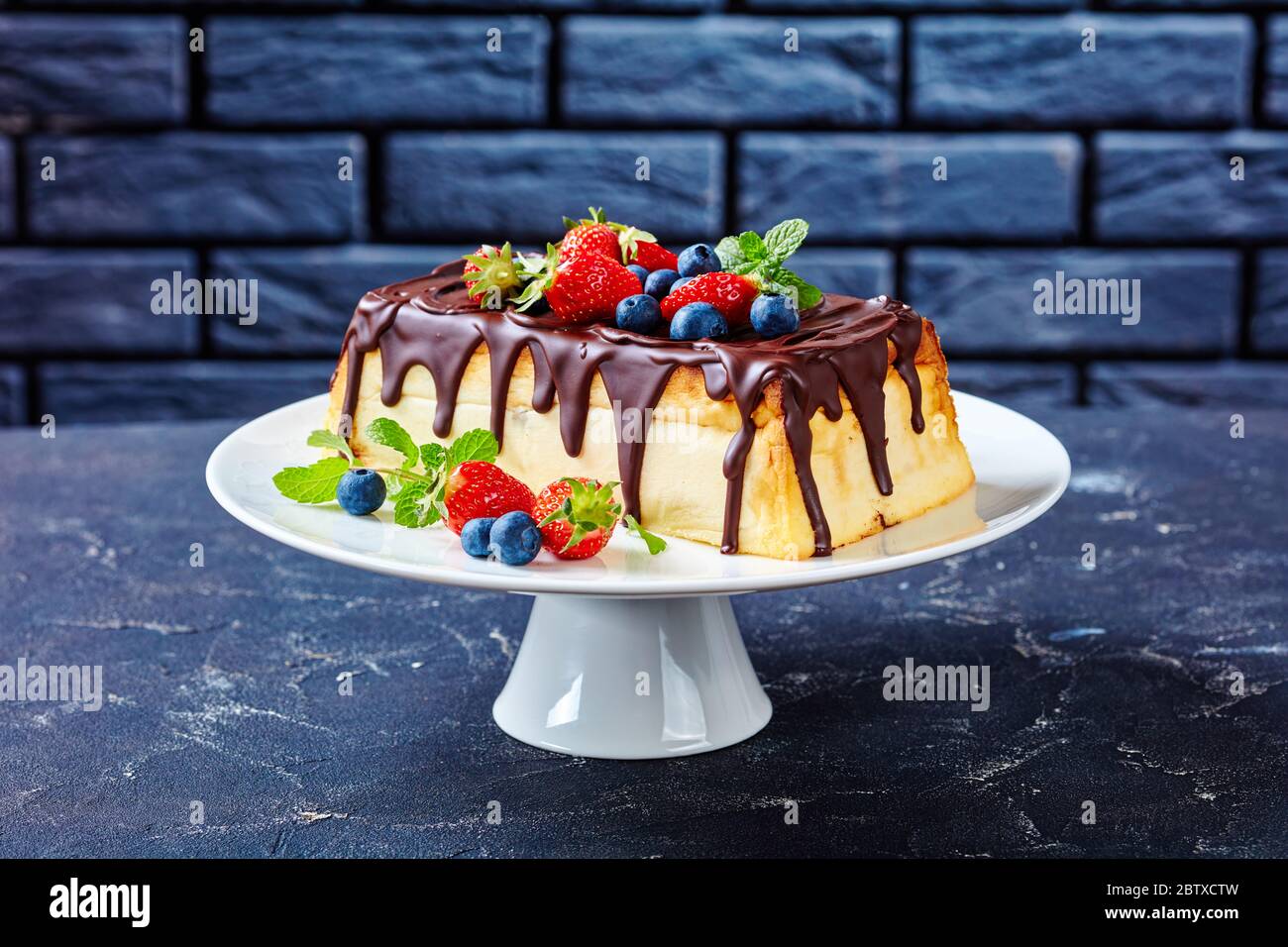 Cheesecake ucraino al formaggio casolare con cioccolato fondente con mirtilli freschi, fragole, foglie di menta su un piedistallo torta bianca su un conc scuro Foto Stock