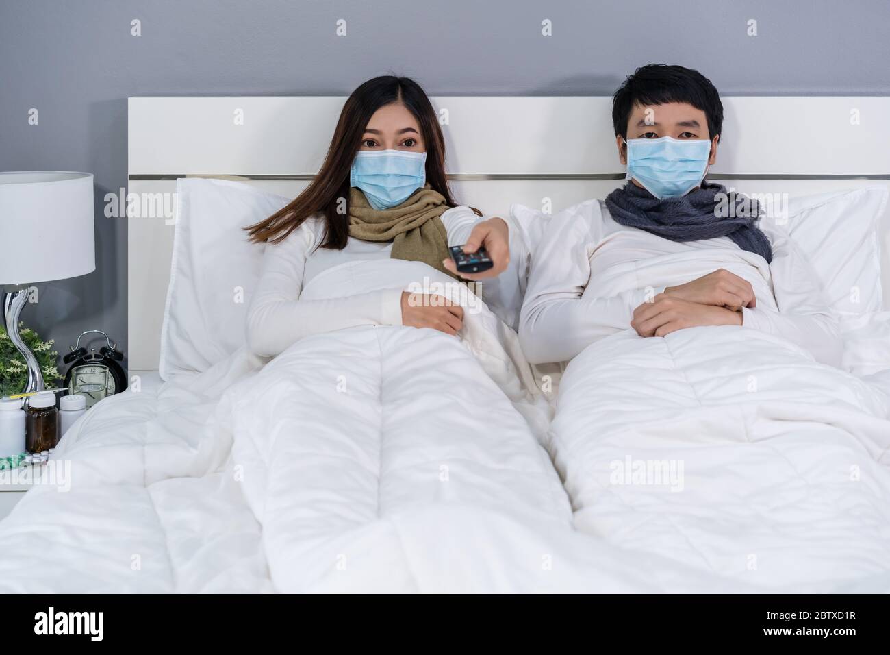 coppia sposata in maschere mediche che guardano la tv su un letto, protezione dal coronavirus (covid-19) concetto pandemico. Foto Stock