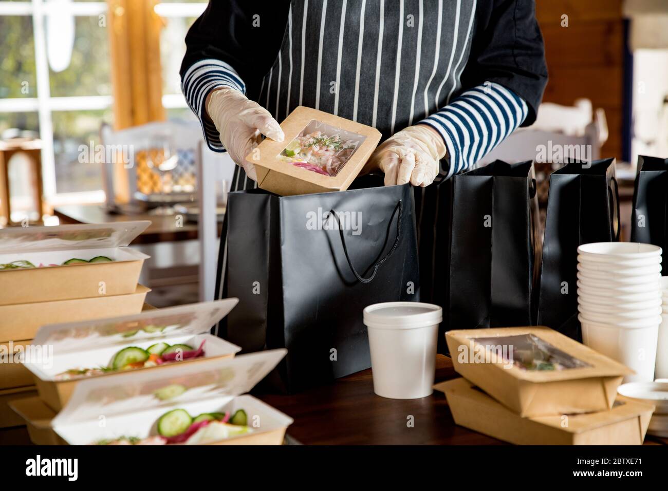Operaio del ristorante che indossa maschera protettiva e guanti che imballano il cibo in scatola take away. Servizi di consegna di cibo e shopping di cibo senza contatto in linea. Foto Stock