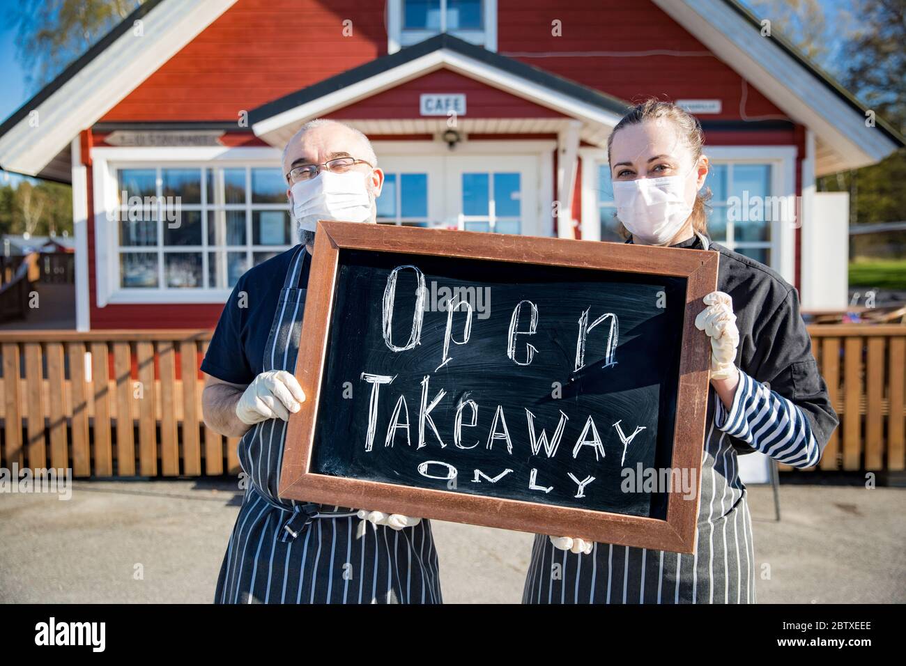 Una coppia di mezza età che indossa maschere protettive in piedi davanti al ristorante e che tiene un cartello solo Takeaway. Piccola impresa durante il coronavirus Foto Stock