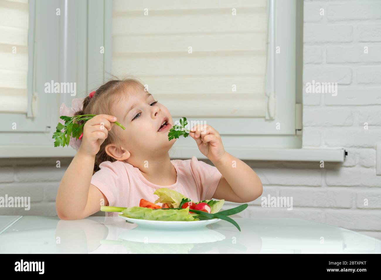 4 anni ragazza seduta in cucina a tavola e mangiando verdure, nelle mani di un rametto di prezzemolo. regole nutrizionali prescolare Foto Stock