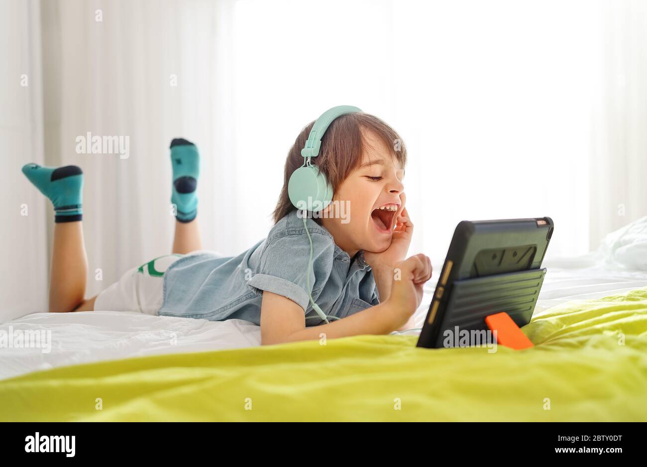 Un ragazzo sorridente seduto a letto e che gioca su un tablet digitale a casa. Bambino in camera da letto indossando le cuffie e utilizzando dispositivi intelligenti divertendosi Foto Stock
