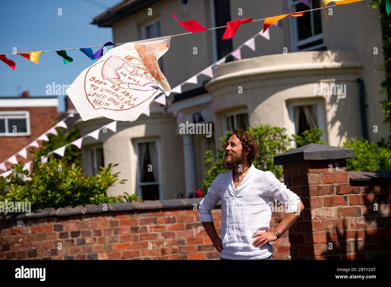 L'artista Luke Perry su Duncombe Street a Wollaston, dove ha appeso bunting con messaggi di sostegno per i vicini e i lavoratori chiave, per creare un corridoio di 500 metri di colore attraverso il villaggio delle West Midlands. Perry ha chiesto ai residenti e ai negozianti locali di presentare disegni e messaggi, che poi dipinse a mano e cucì su conchettando con l'aiuto di volontari. Foto Stock