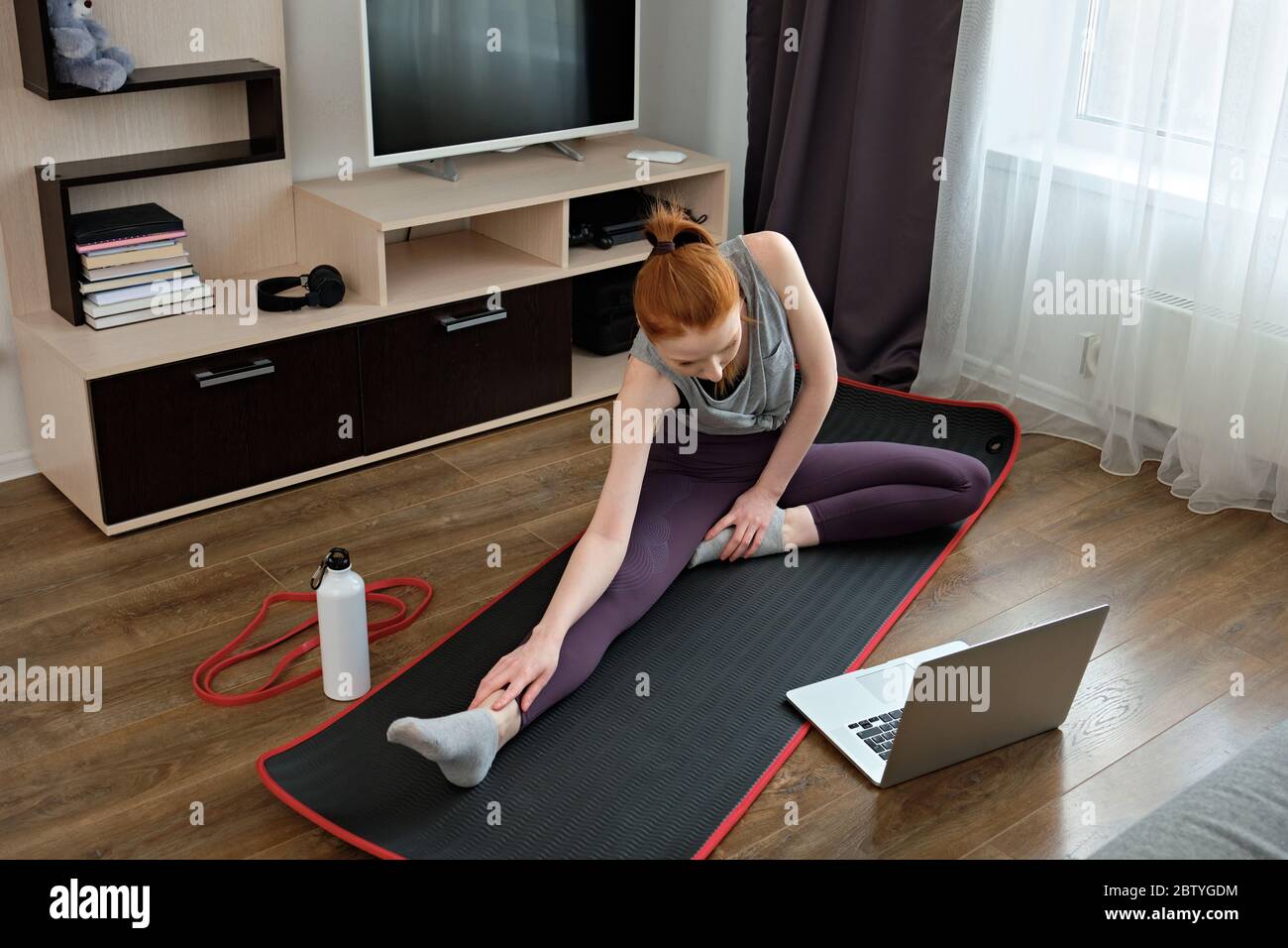 Ragazza rossa in abbigliamento sportivo che si allunga a casa, seduto di fronte a un laptop, telaio dall'alto. Foto Stock