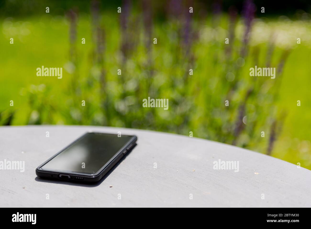 Spento smartphone sdraiato su un tavolo da giardino dipinto di grigio con fiori in fiore sullo sfondo, mettere a fuoco sullo smartphone Foto Stock