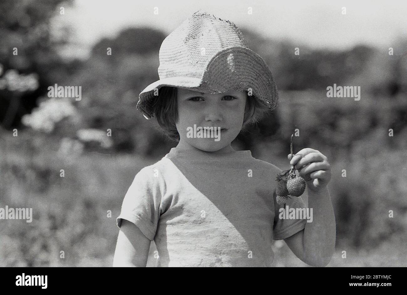 1987, estate e una bambina con un cappello di paglia che mostra una fragola che ha appena scelto, Inghilterra, Regno Unito. Foto Stock