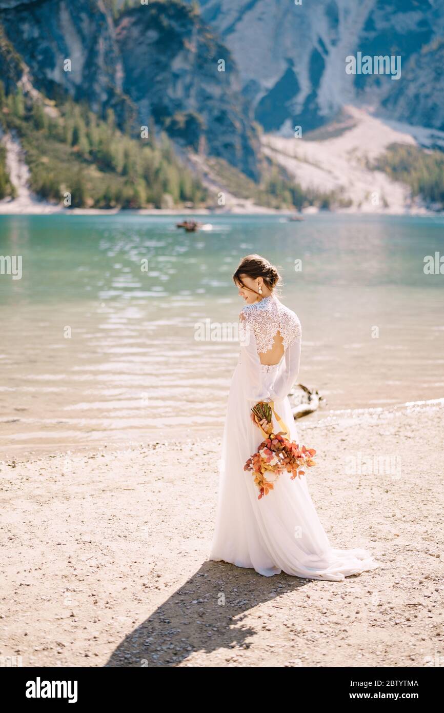 Bella sposa in abito bianco con maniche e pizzo, con bouquet giallo autunnale di fiori secchi e rose di pony, sul Lago di Braies in Italia Foto Stock