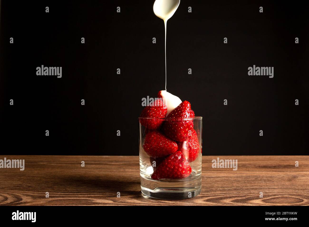 Fragole in un bicchiere con un cucchiaio e latte condensato su fondo scuro Foto Stock