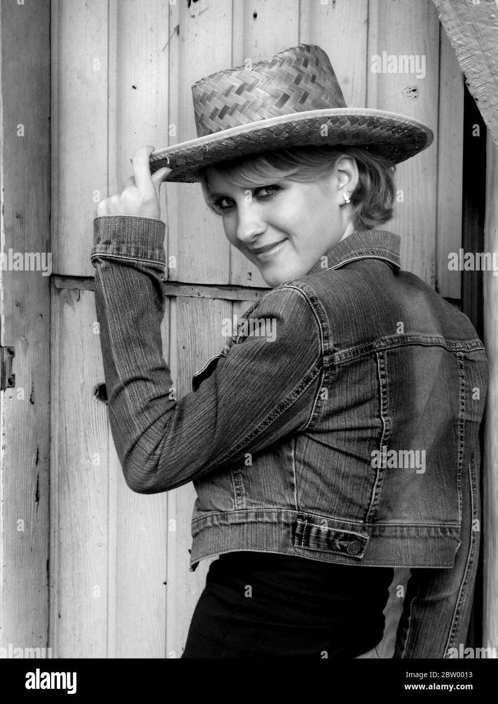 Guardaroba bianco e nero di giovane donna con jeans e giacca in denim. Sta ribaltando il brim del suo cappello di paglia e grinning. Foto Stock