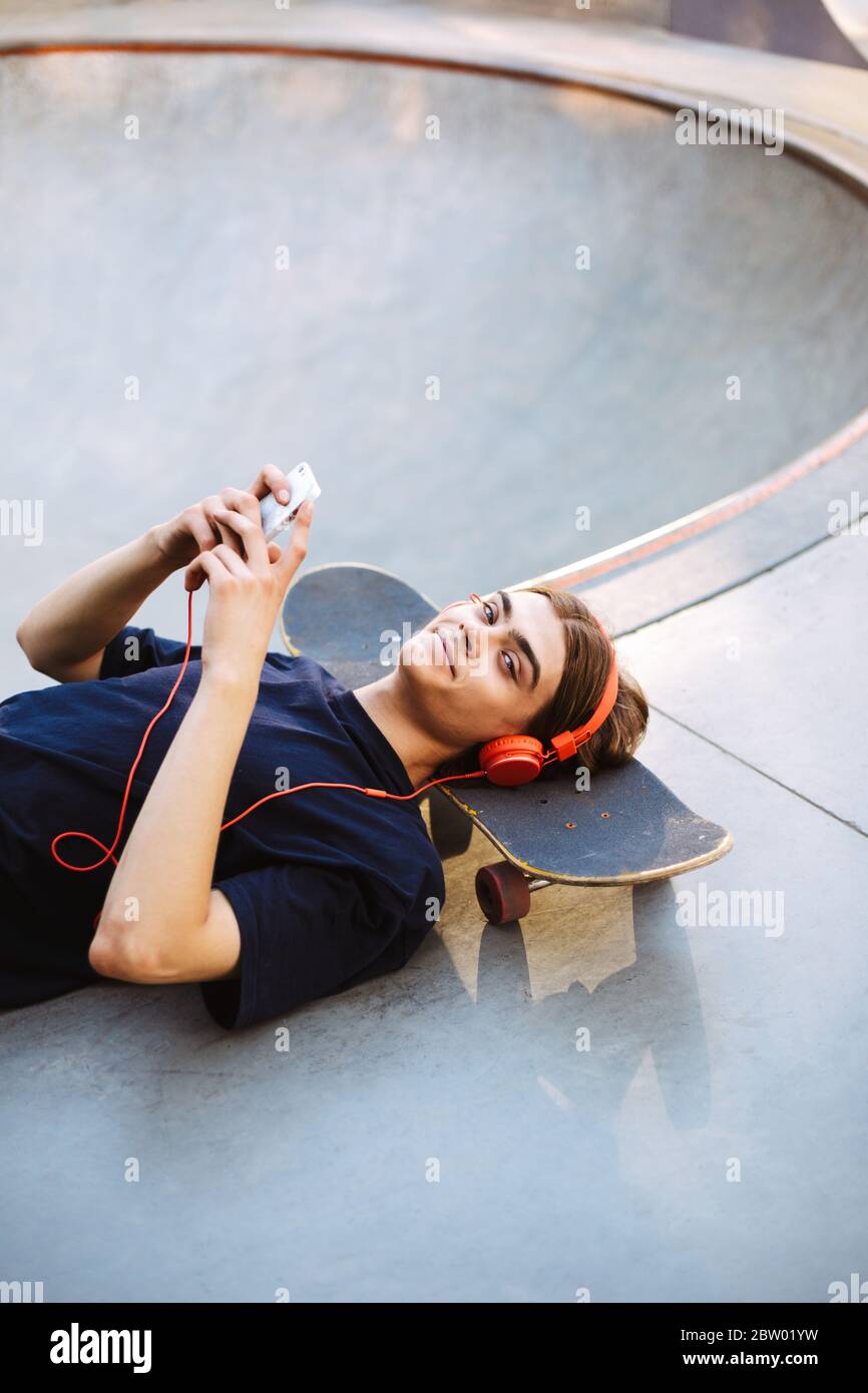 Ragazzo in cuffie arancioni sdraiato sullo skateboard con il cellulare in mano mentre felicemente guardando in macchina fotografica al parco skatepark isolato Foto Stock