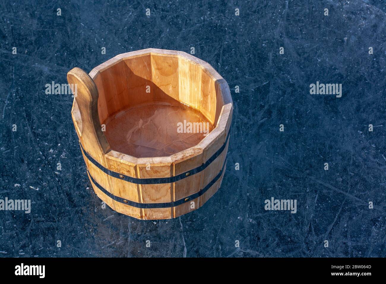 Una vasca di legno con ghiaccio all'interno è in piedi sul ghiaccio con crepe. Gli anelli di serraggio in ferro collegano le parti della vasca. Soleggiata. Orizzontale. Foto Stock