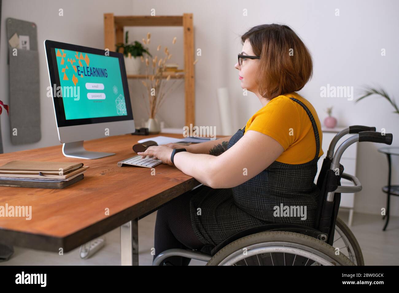 Donna giovane e seria con disabilità in occhiali seduti in sedia a rotelle e entrando nel login mentre si prepara ad iniziare la lezione di e-learning a casa Foto Stock