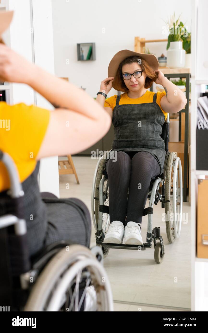 Bella donna porta in occhiali seduta in sedia a rotelle davanti allo specchio e indossare un bel cappello prima di passeggiare Foto Stock