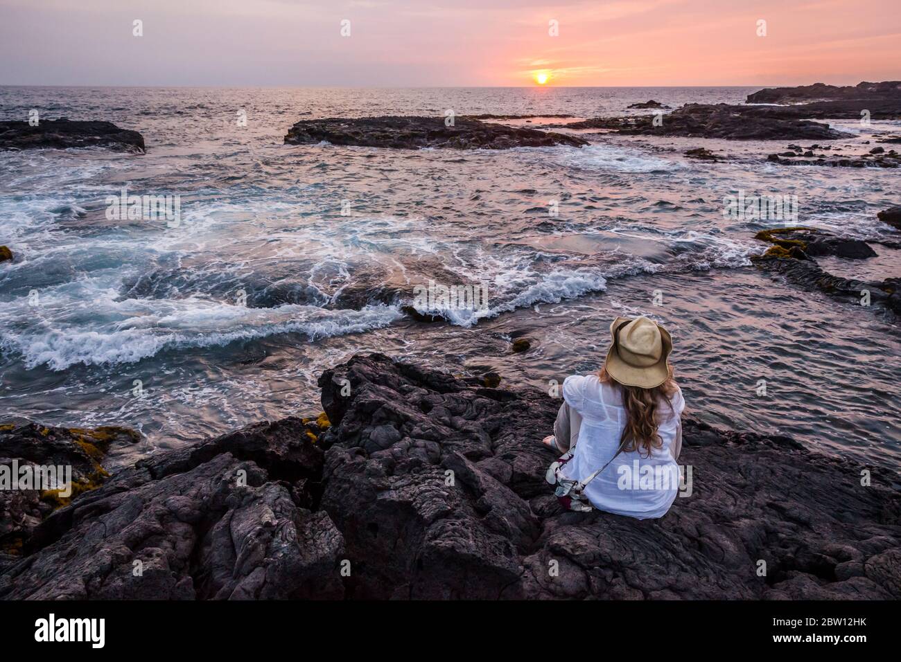 Donna siede su vecchie rocce laviche al bordo delle acque guardando il tramonto, Wawaloli Beach Park, Kailua-Kona, Hawaii, USA. Foto Stock