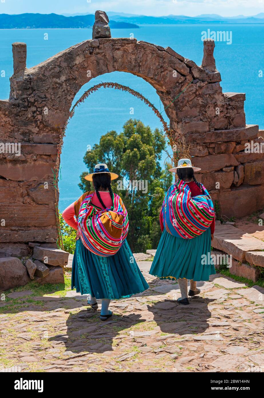 Due donne indigene peruviane di Quechua in abiti tradizionali che camminano attraverso l'arco dei governanti sull'isola di Taquile con il lago Titicaca, Perù. Foto Stock