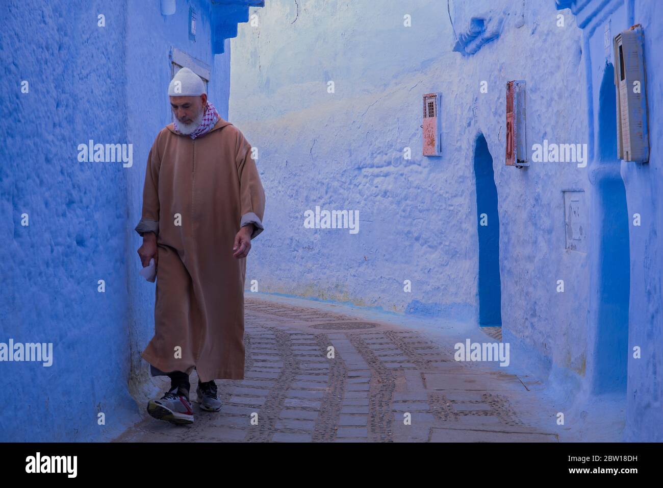 Un marocchino locale in abiti tradizionali cammina lungo una stretta vicolo acciottolato nella parte della città vecchia di Chefchaouen, Marocco. Foto Stock
