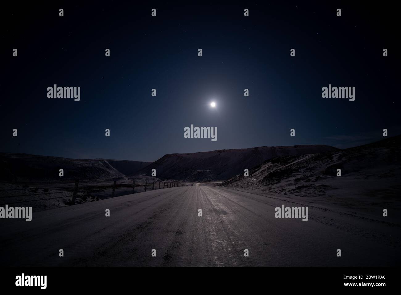 Vuota strada ghiacciata in una remota posizione in Islanda illuminata solo da una luna e con un cielo stellato in una notte limpida. Foto Stock