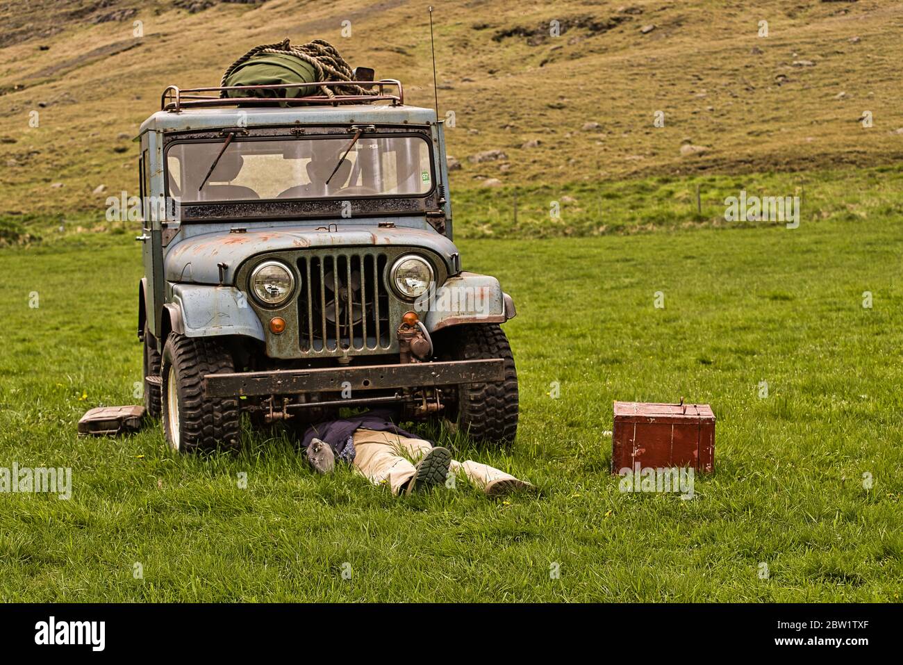 Meccanico o conducente sotto un veicolo fuori strada rotto, vecchio e fatiscente posto su un prato verde. Cassetta degli attrezzi rossa metallica. Foto Stock