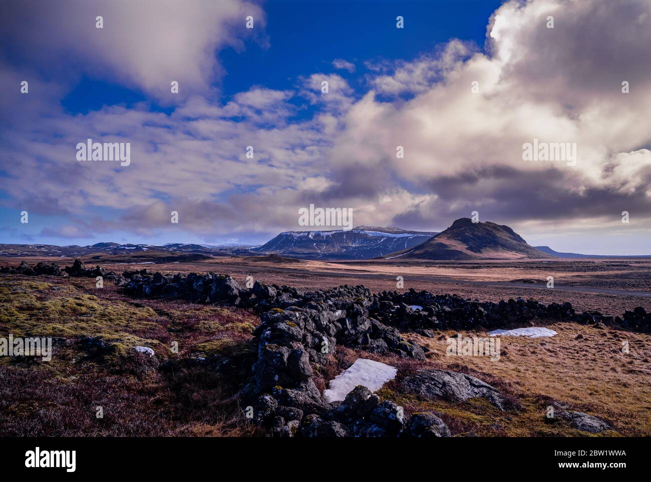 Un vecchio muro di roccia nel paesaggio roccioso dell'Islanda occidentale con una grande montagna, una catena di montagna coperta di neve, e un cielo nuvoloso sullo sfondo. Foto Stock