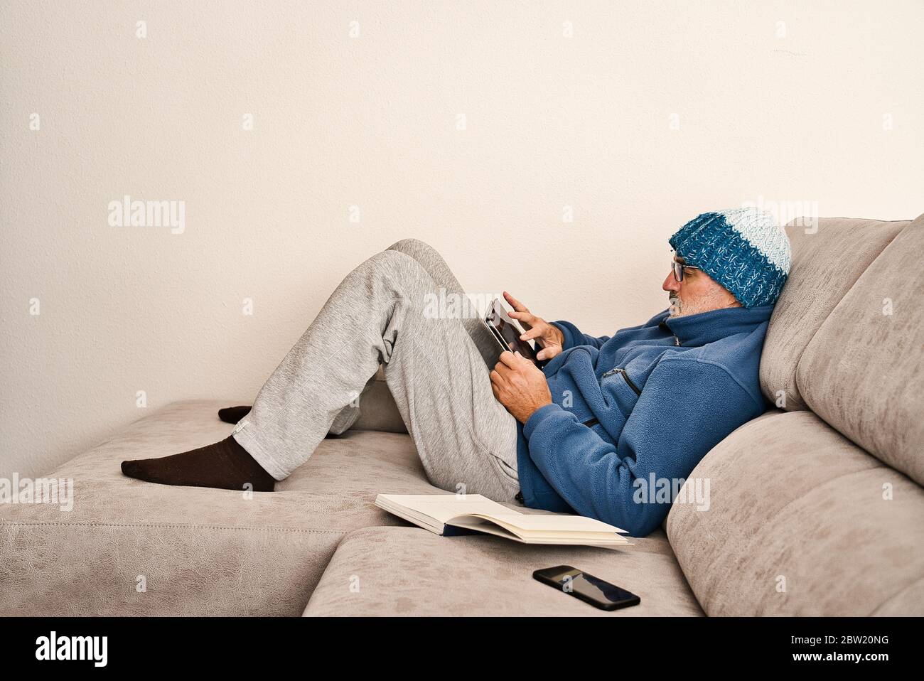 Un adulto con bicchieri, una barba corta e un cappuccio di lana riposa comodamente sul divano grigio di una casa moderna e minimalista. Collegamento e controllo Foto Stock