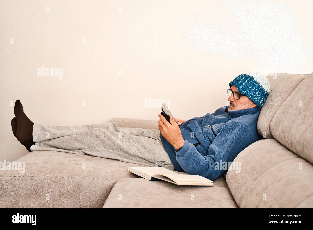 Un adulto con bicchieri, una barba corta e un cappuccio di lana riposa comodamente sul divano grigio di una casa moderna e minimalista. Collegamento e controllo Foto Stock