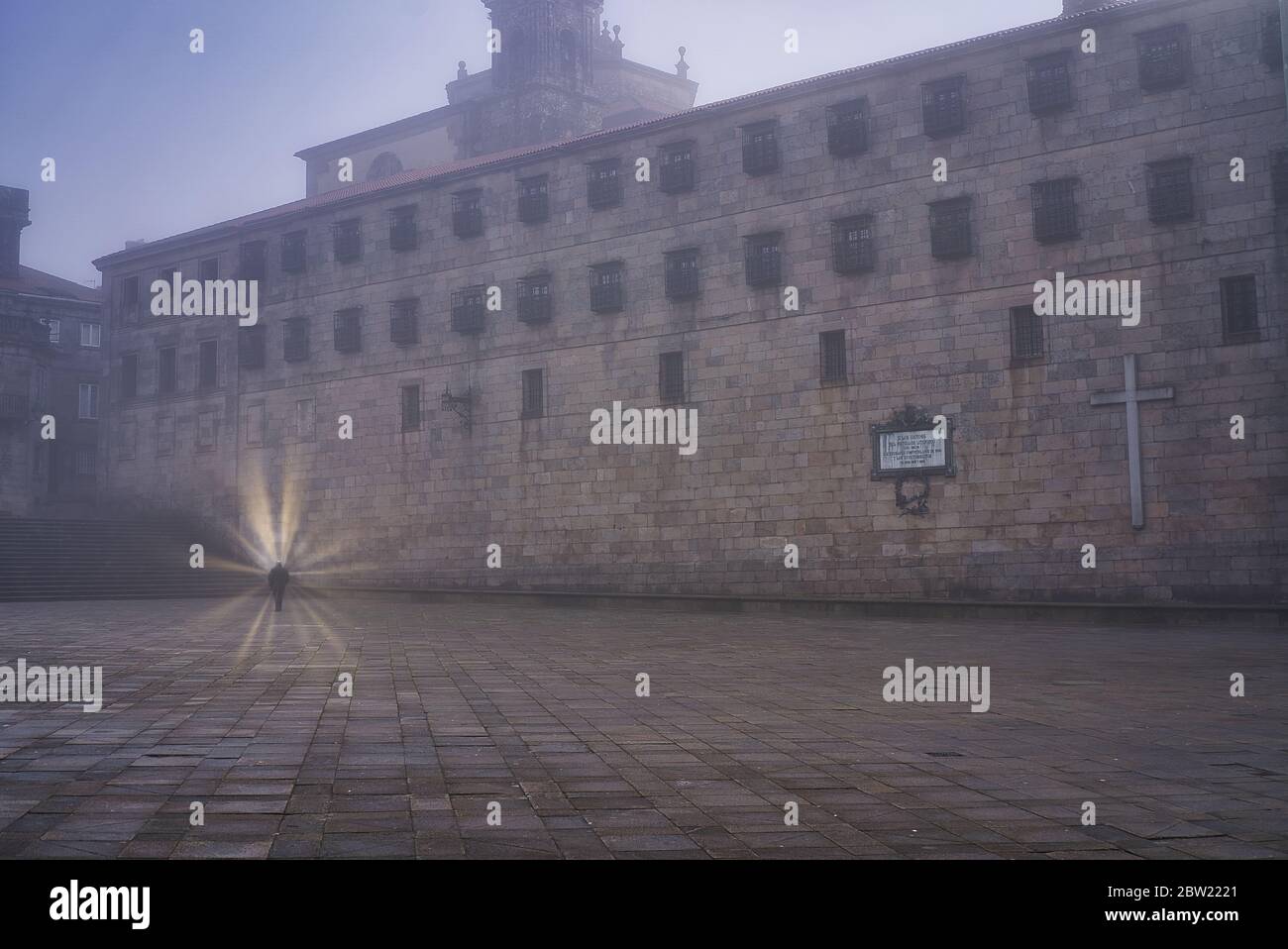 Una persona circondata da un'aura divina che cammina da sola per le strade di un'antica città accanto a un monastero cattolico in un giorno d'autunno con nebbia fitta Foto Stock
