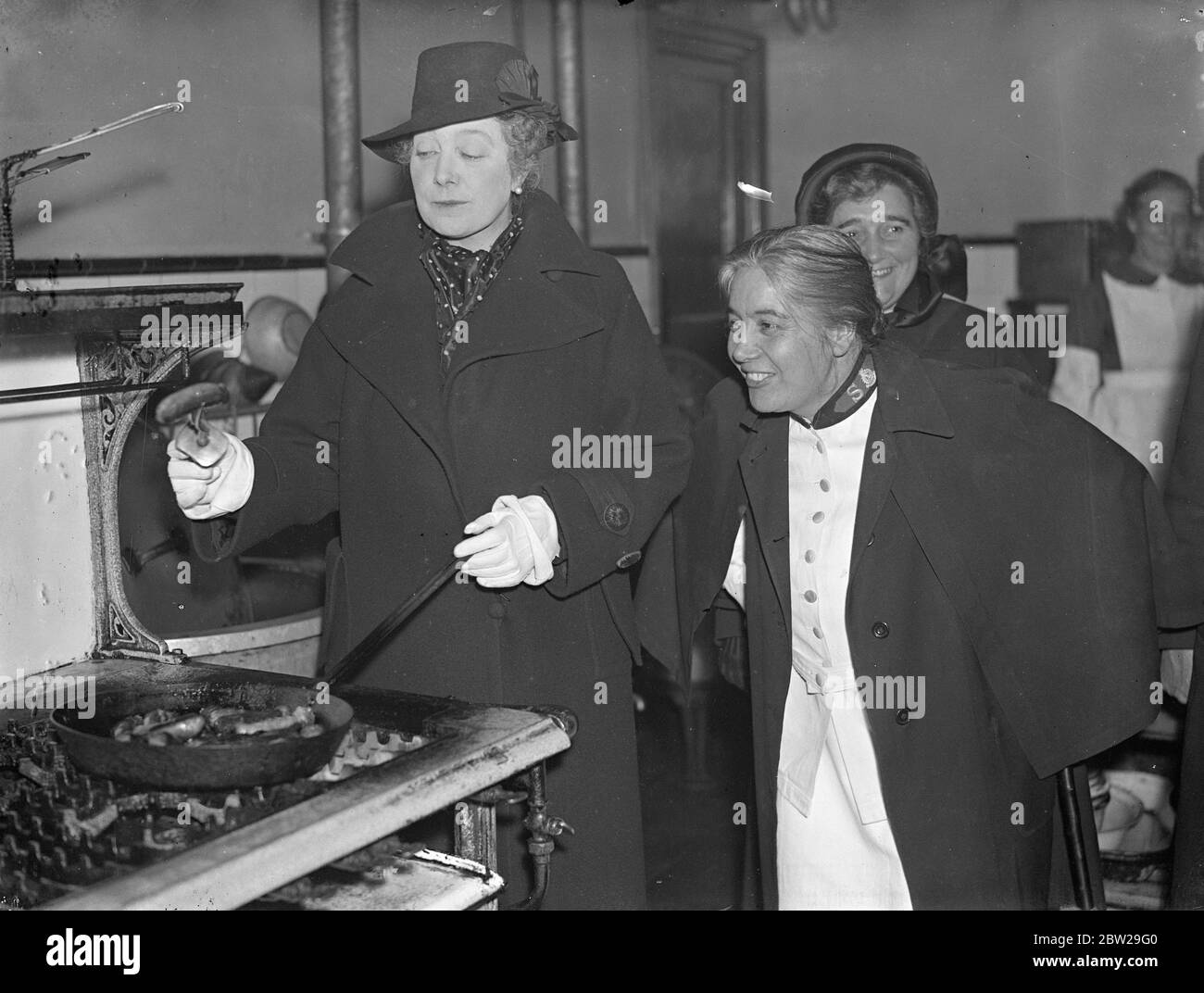 Premier's moglie, salsicce patatine fritte, durante la visita di East End ostello. La signora Neville Chamberlain, moglie del Premier, salumi fritti nelle cucine dell'ostello quando ha visitato l'ospedale femminile dell'esercito della salvezza a Finch Street, Whitechapel. Mostra fotografica, la signora Chamberlain tiene in mano una salsiccia fritta per l'ispezione. Accanto a lei è Dennett maggiore, matron dell'ostello. 15 novembre 1937 Foto Stock