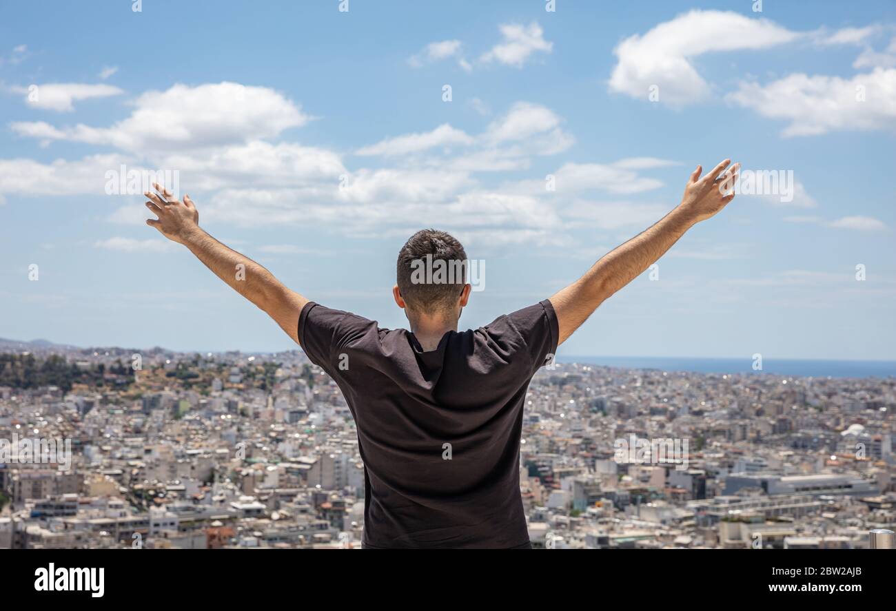 Uomo dietro con braccia sollevate. Vista posteriore per giovani adulti da soli con mani aperte che guardano la città sotto, sfondo blu del cielo. Stile di vita sano, fre Foto Stock