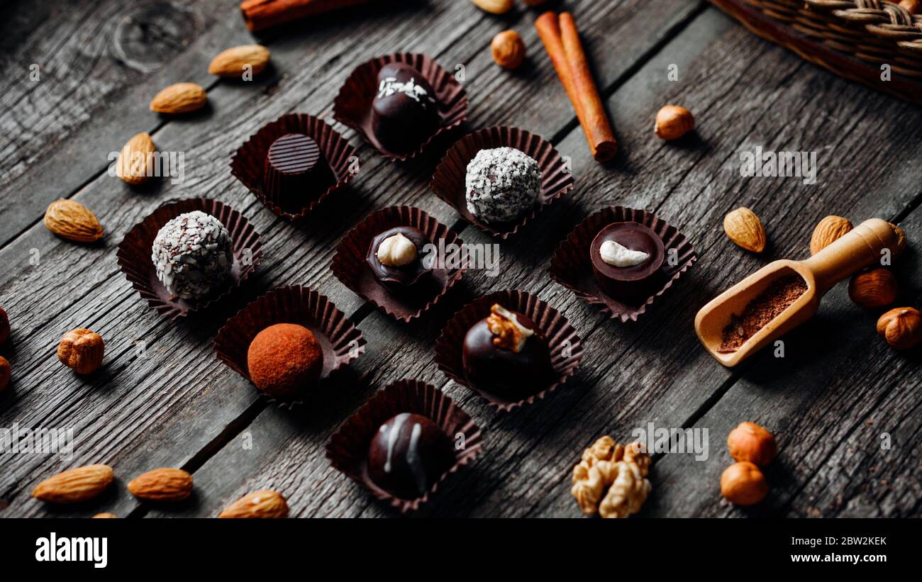 Nove caramelle di cioccolato fatte a mano all'interno di involucri caramelle su sfondo di legno vintage. Noci, cannella, mandorla, nocciole intorno. Foto Stock