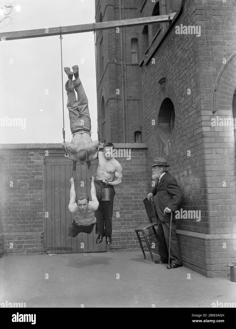 Circus acrobat uomini forti che praticano una routine in un cortile di mattoni . Tre uomini equilibrati su anelli di sospensione, mentre un Signore che trasporta sedia e canna guarda sopra Foto Stock