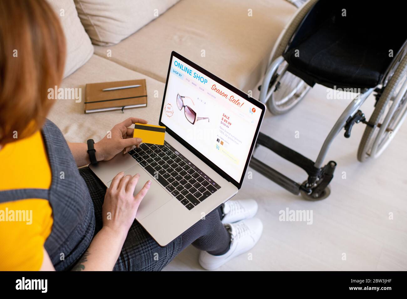 Vista a spalla di disabili donna seduta sul divano e utilizzando il computer portatile mentre si sceglie occhiali da sole in negozio di Internet Foto Stock