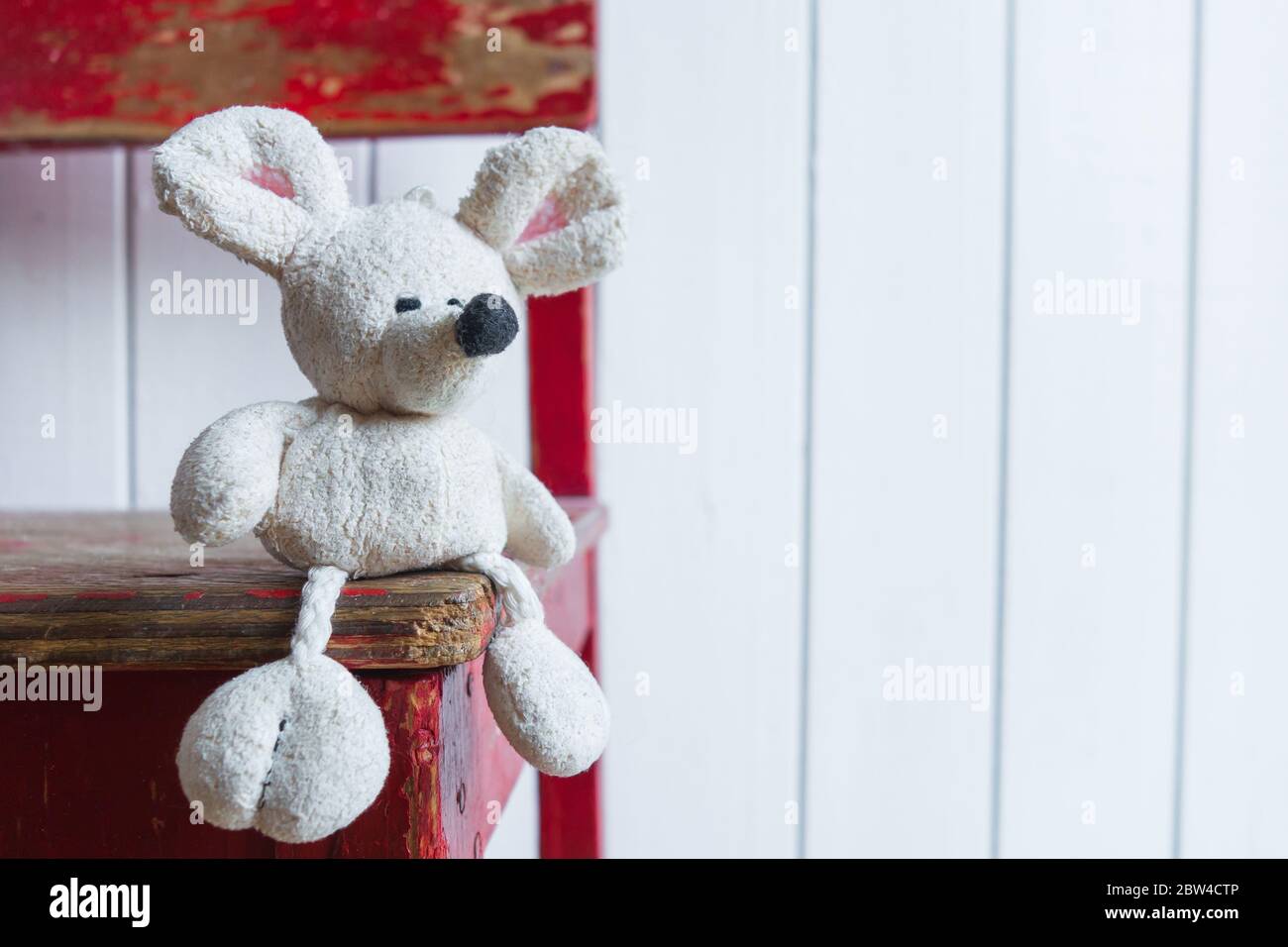 Un vecchio mouse bianco si siede su una vecchia sedia rossa sullo sfondo di una parete bianca. Concetto di solitudine nell'infanzia e nell'età avanzata. Foto Stock
