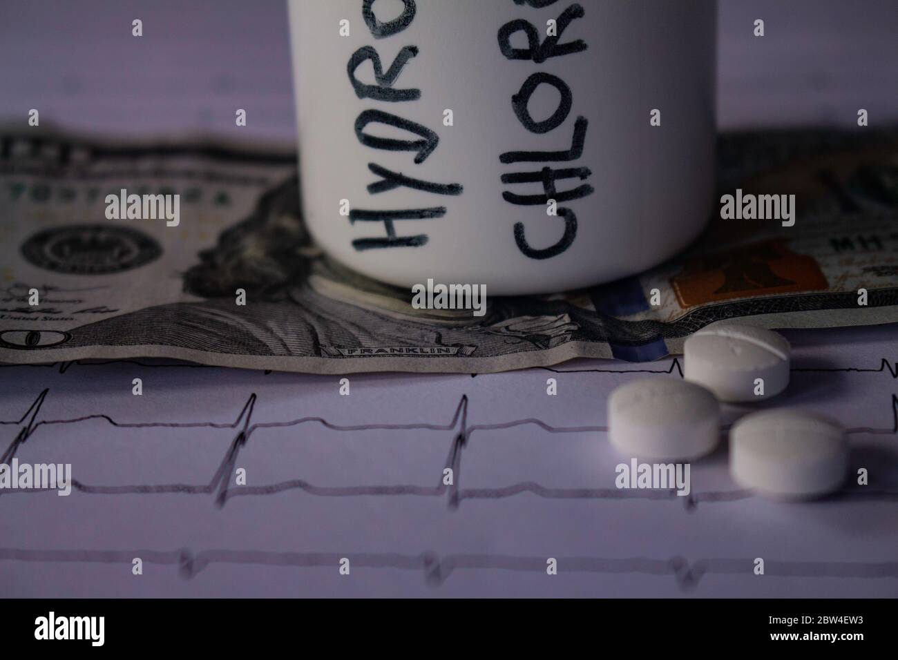 Pillole su un elettrocardiogramma. Cento dollari di bollette su un EKG. Contenitore bianco con 'idrossiclorochina' scritto sul lato. Concetto di costo elevato di Foto Stock