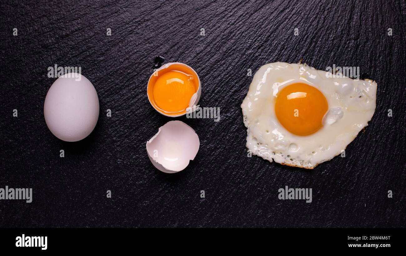 sulla pietra ardesia nera, con vista dall'alto, in sequenza, un uovo bianco, un tuorlo d'uovo nel guscio e un uovo fritto. Vita morta Foto Stock