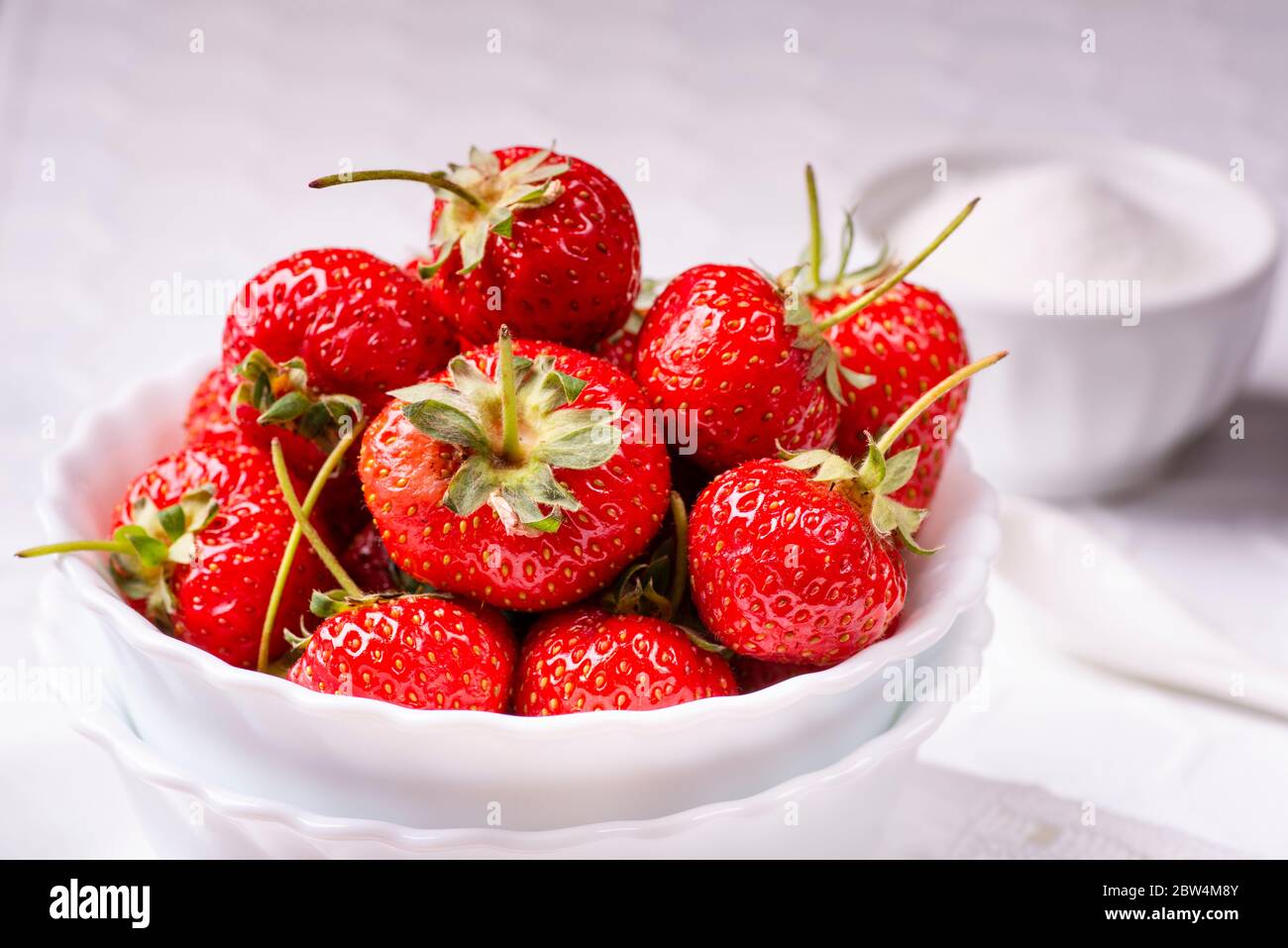 sulla tovaglia bianca testurizzata, in primo piano, una ciotola bianca in ceramica con fragole rosse mature e brillanti Foto Stock