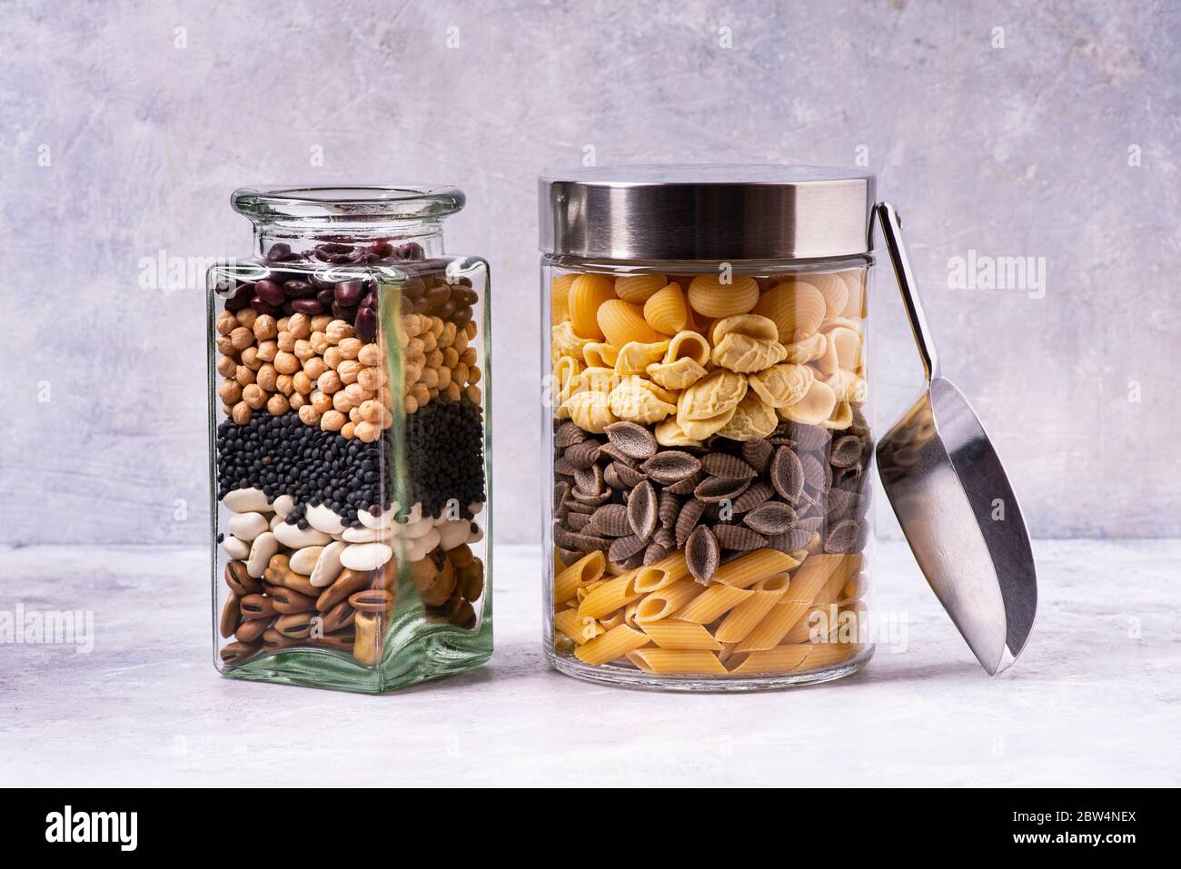 Su uno sfondo grigio testurizzato, varietà di pasta italiana e legumi secchi in vasi di vetro in primo piano. Dieta mediterranea Foto Stock
