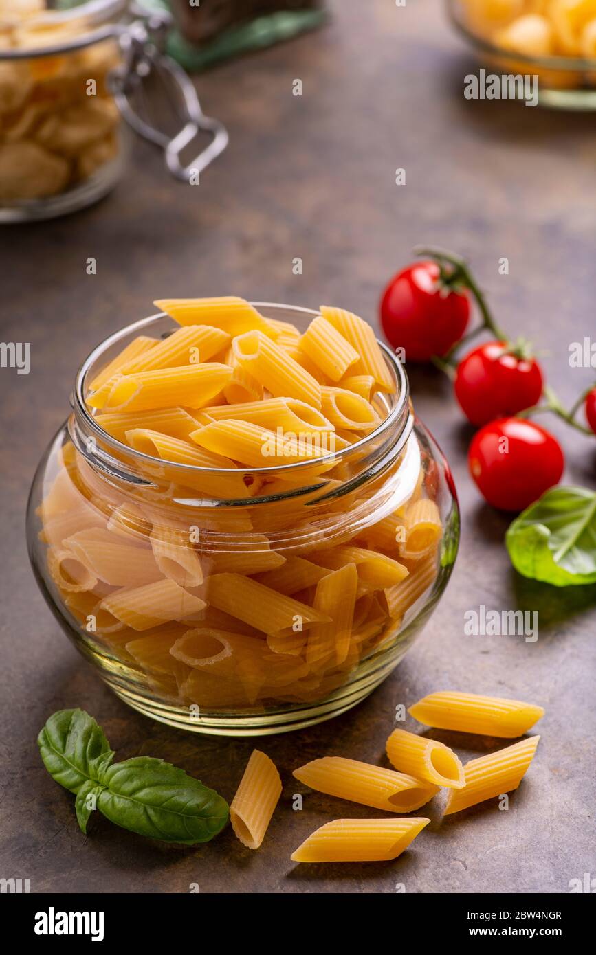 In primo piano in un vaso di vetro trasparente, pasta italiana cruda, sullo sfondo mazzo di pomodori ciliegini e foglie di basilico fresco. Foto Stock