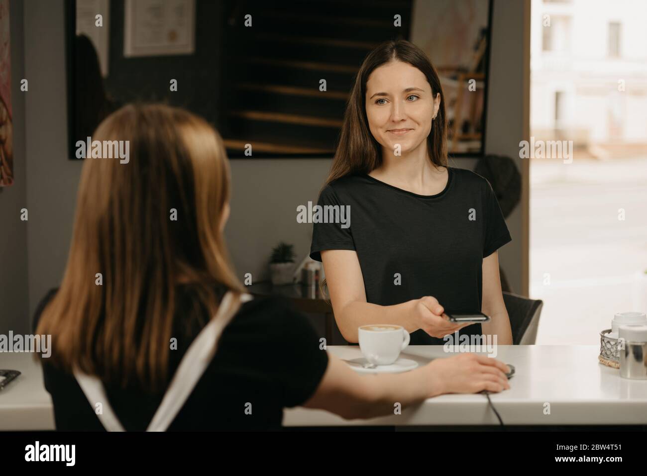 Una ragazza felice con i capelli lunghi sorridente e pagando per il suo caffè con uno smartphone da contactless PAY PASS tecnologia in un caffè. Una barista femminile di brunette Foto Stock