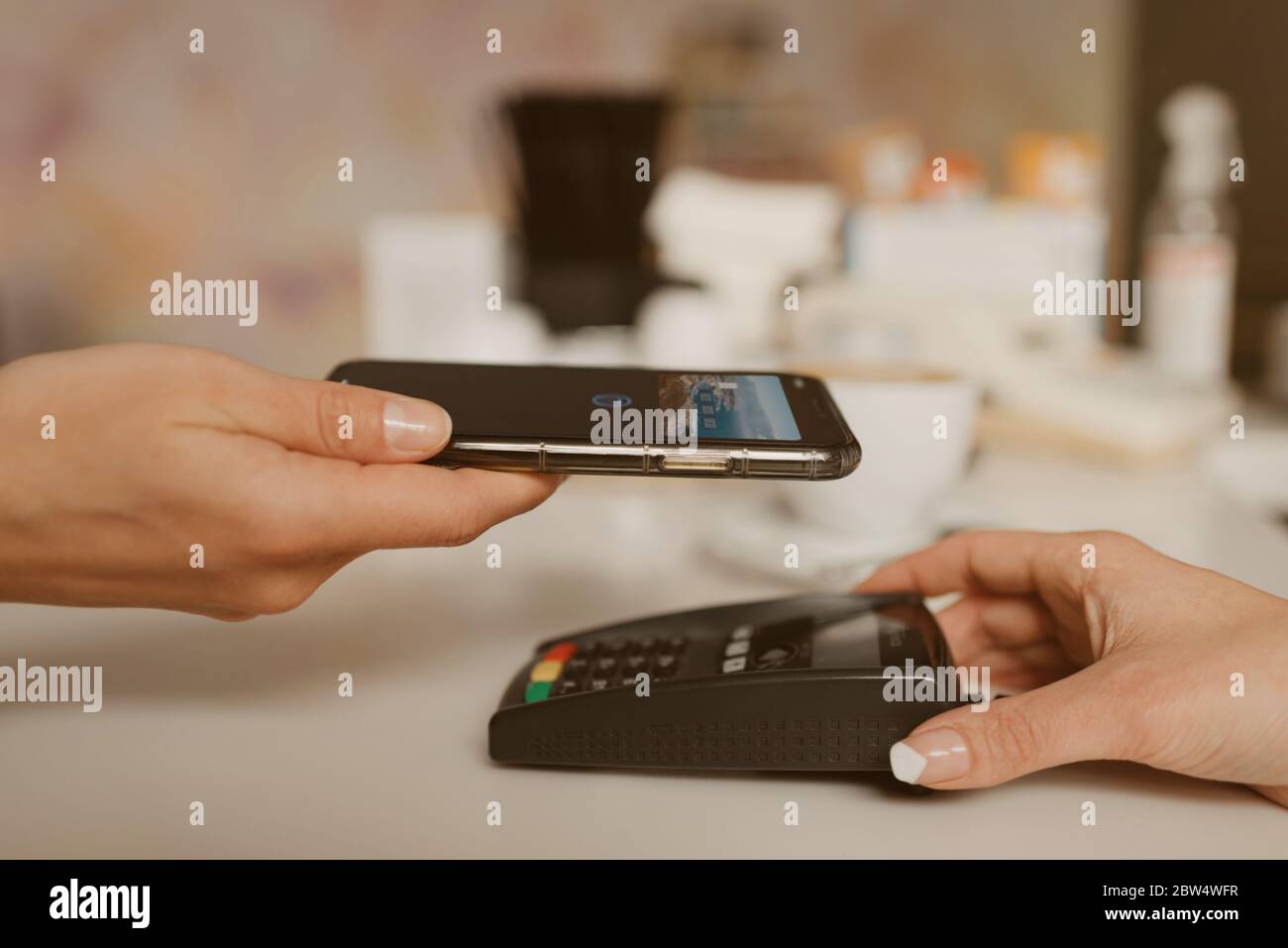 Una foto ravvicinata di uno smartphone che si prepara a pagare un latte con la tecnologia PAY PASS senza contatto in un bar. Un barista femminile tiene fuori un terminale per Foto Stock