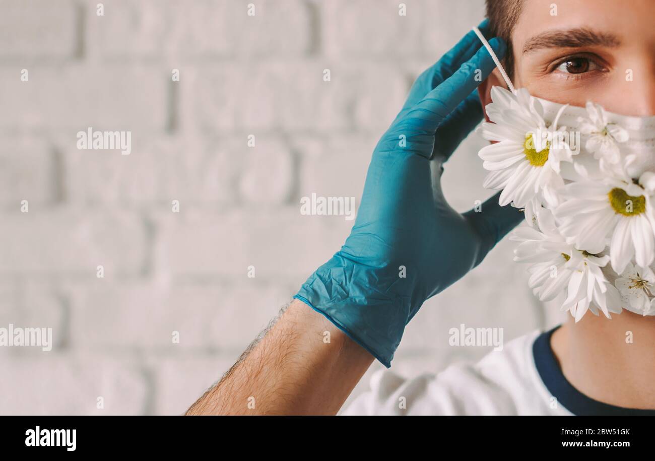 Closeup medico felice maschio in lattice guanti protettivi indossare, togliendo maschera medica viso con fiori margherita. Giovane uomo ha messo o rimuovere prote estate Foto Stock