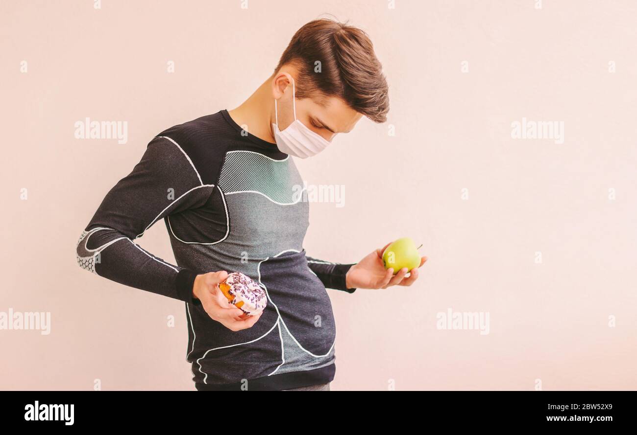 Divertente sport uomo in maschera medica viso con pancia grande lotta tra frutta sana e cibo spazzatura. Grasso giovane uomo fitness in maschera protettiva tenere swe Foto Stock