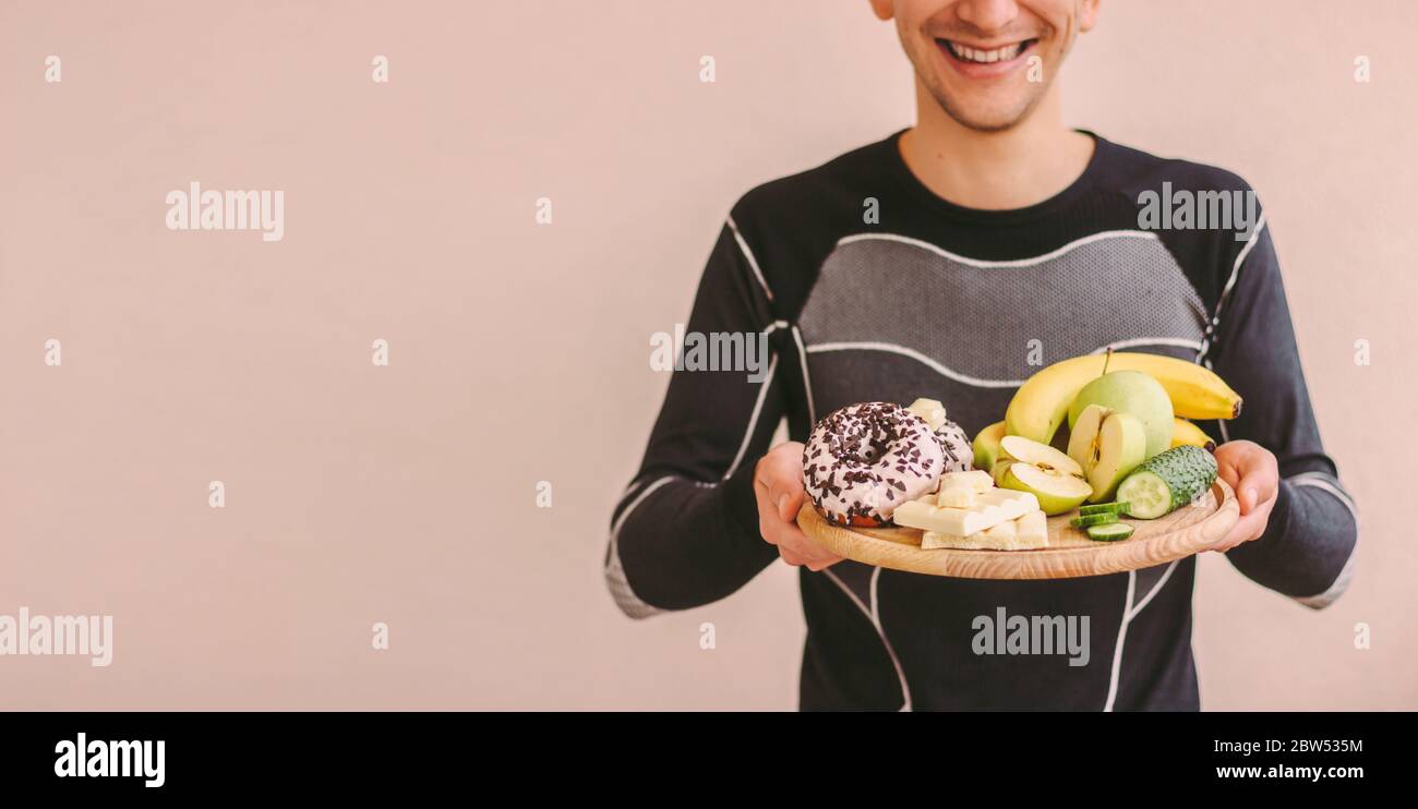 Banner closeup di felice uomo sportivo tenere tavola di legno con frutta fresca, verdure, cioccolatini, ciambella. Giovane uomo affamato di fitness scegliendo tra healt Foto Stock