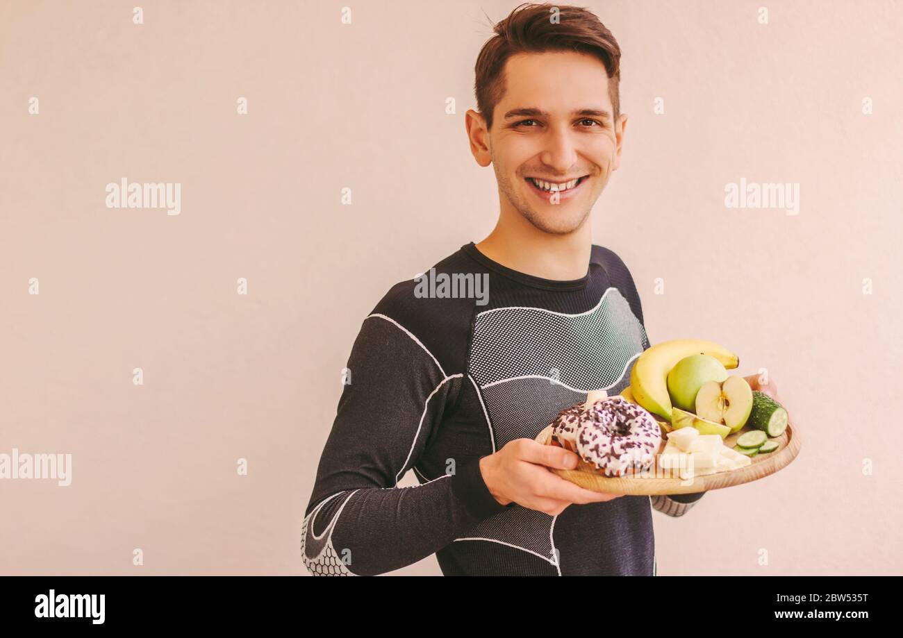 Giovane uomo sportivo che decide cosa mangiare: Frutta sana o zucchero spazzatura snack malsani. Uomo sportivo felice che tiene tavola di legno con mele fresche, banane Foto Stock