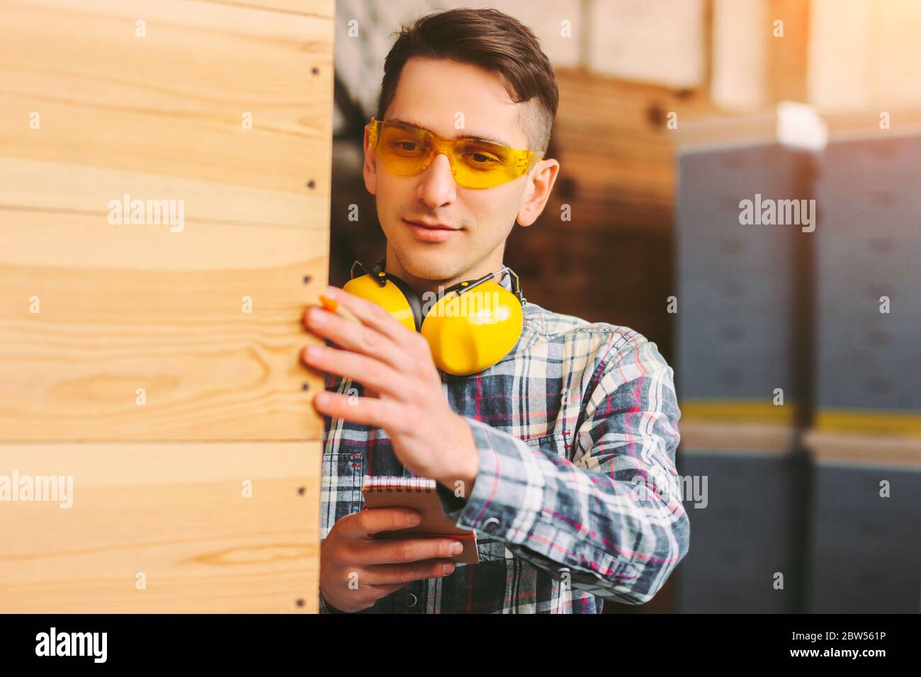 Responsabile magazzino giovane uomo in occhiali protettivi, cuffie contare materiale di produzione legno e sorridente. Operaio di segheria maschile che ispeziona legno pr Foto Stock