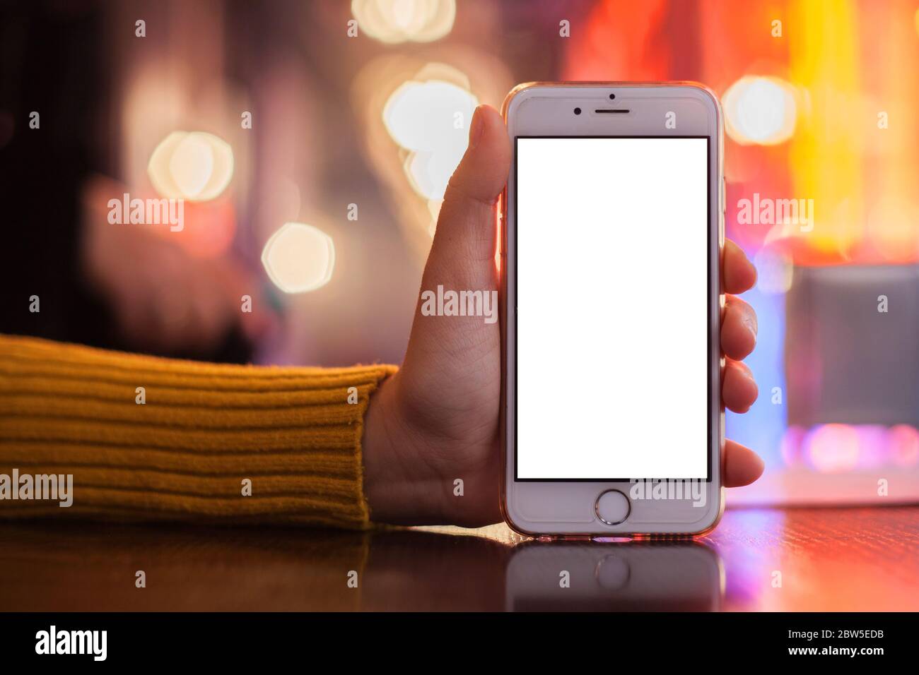 Immagine mockup di una mano che tiene un telefono cellulare bianco con uno schermo bianco vuoto. Sfondo sfocato con bokeh colorato su un tavolo in un bar. Messa a fuoco selettiva Foto Stock